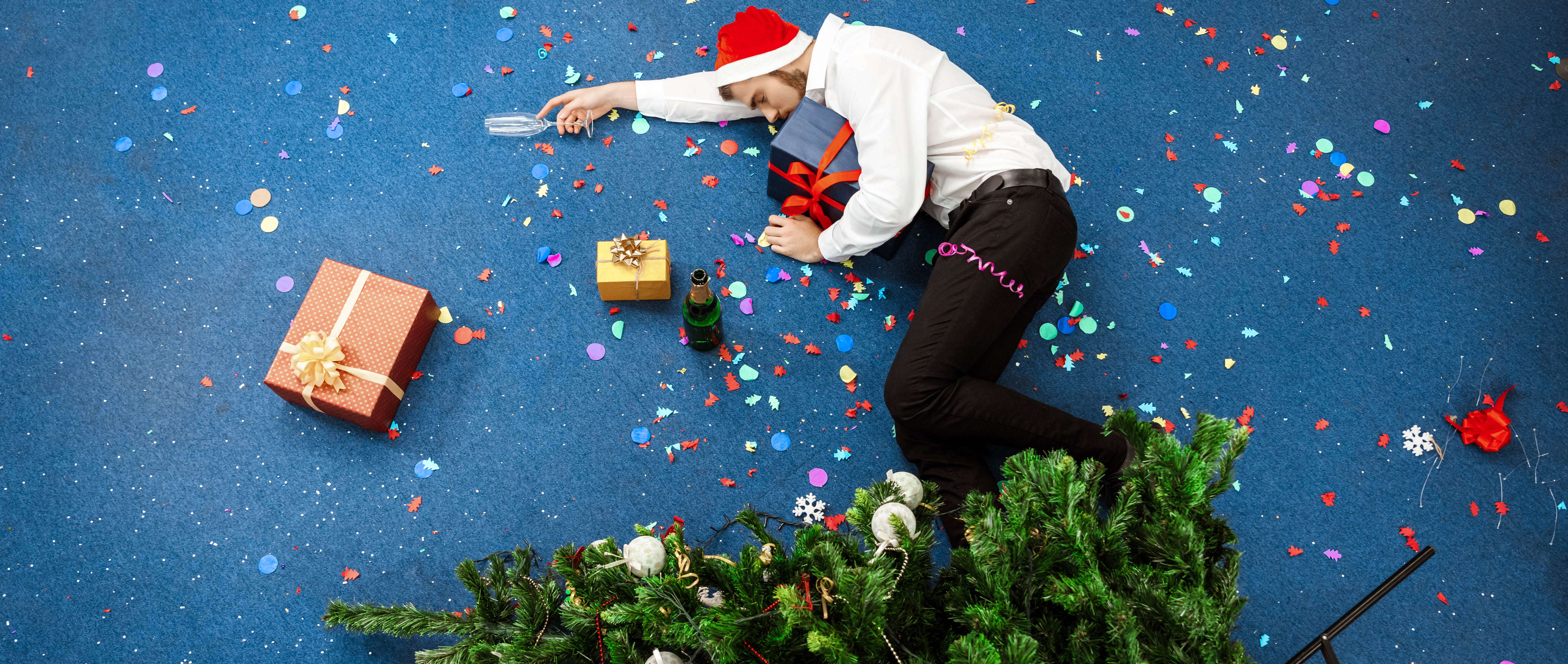 Man asleep on the floor after a holiday party - empty champagne glass in his hand, Santa hat on his head, and Christmas tree tipped down onto his legs Man asleep on the floor after a holiday party - empty champagne glass in his hand, Santa hat on his head, and Christmas tree tipped down onto his legs